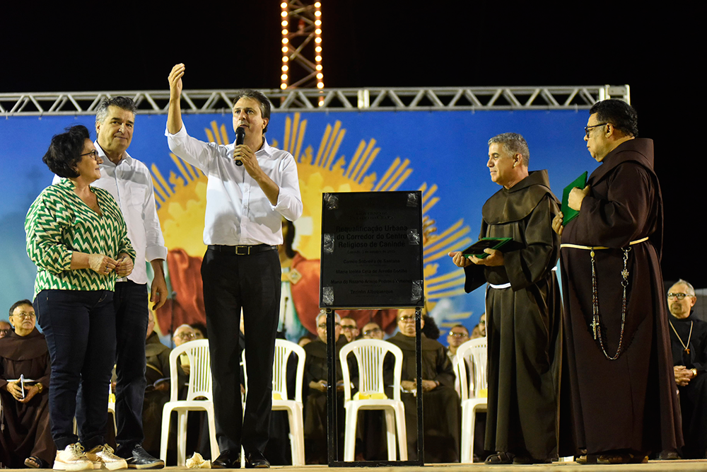 Foto de Davi Pinheiro traz o Governador do Ceará, Camilo Santana sobre o palco, com a mão direita levantada. O governador anuncia os investimentos para cidade de Canindé. Ao lado de Camilo Santana, frades de Canindé.